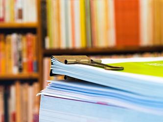 pile of journals and paper records with large paper clip and library shelves behind